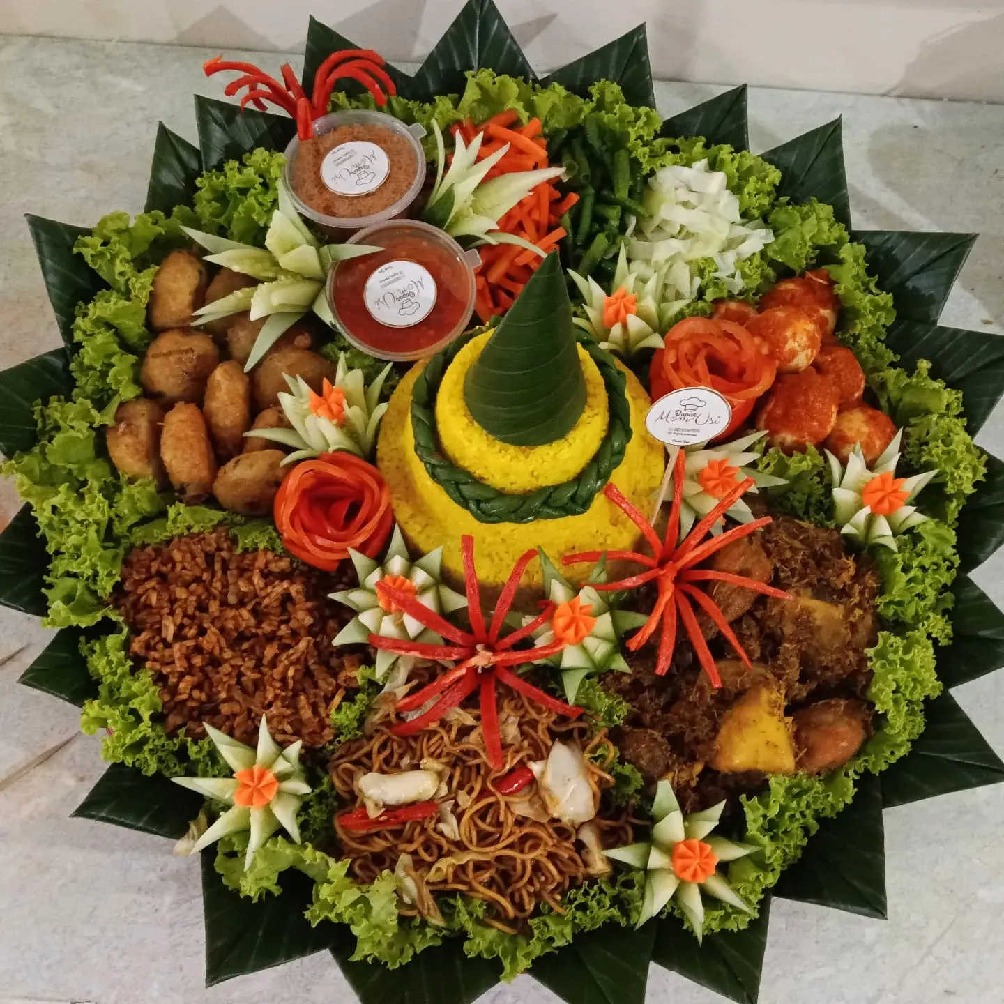 Tumpeng tampah Dapur Mom'Usi tampak atas: nasi kuning bertingkat dikelilingi mi goreng, orek tempe, rendang, perkedel, telur balado, dan hiasan ukir sayuran di atas alas daun pisang